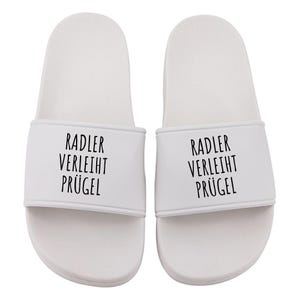 Könnte beinhalten: Ein Paar weiße Badesandalen. Jede Sandale hat einen weißen Riemen mit dem schwarzen Text "RADLER VERLEIHT PRÜGEL". Die Sandalen sind für den lässigen Gebrauch konzipiert und eignen sich für den Innen- und Außenbereich.
