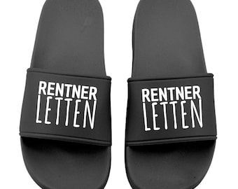 Könnte beinhalten: Ein Paar schwarze Badesandalen. Auf jedem Riemen steht in weißer Schrift "RENTNER LETTEN". Die Sandalen sind für den lässigen Gebrauch konzipiert und für den Innen- und Außenbereich geeignet.