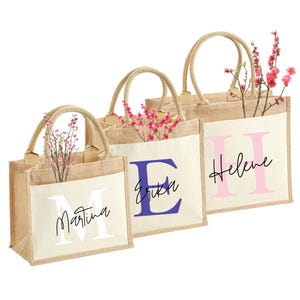Könnte beinhalten: Drei Jute-Tragetaschen mit cremefarbenen Fronten, jede mit einem anderen Anfangsbuchstaben und Namen in einer Kontrastfarbe. Die Taschen haben beige Griffe und sind mit rosa Blumen gefüllt. Die Namen sind Martina, Erika und Helene.
