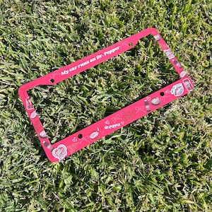May include: A pink license plate frame with the text "My car runs on Dr. Pepper." The frame is decorated with Dr. Pepper logos, hearts, and other graphics. The frame is made of metal and has pre-drilled holes for mounting.