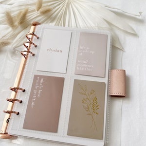 Peut inclure: Un classeur en plastique transparent avec quatre cartes à l'intérieur. Les cartes sont beiges et ont du texte blanc. Le texte sur les cartes est "elysian", "life is made up of small moments like this", "inhale love exhale gratitude", et un motif floral. Le classeur a un mécanisme d'anneaux en or rose.