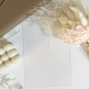 May include: A white envelope with a triangular flap and a row of holes down the side. The envelope is on a white surface with a white and pink flower bouquet and a white candle in the background.