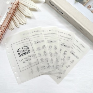 May include: A set of clear plastic recipe cards with black text and icons. The cards are designed for a recipe binder and include sections for recipe name, difficulty level, ingredients, and instructions. The cards feature a variety of food icons, including avocado, salmon, onion, garlic, and more.