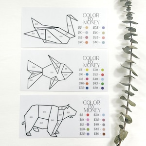 May include: Three white cards featuring origami animal designs: a crane, a fish, and a bear. Each design is divided into sections labelled with dollar amounts. The text "COLOR BY MONEY" appears on each card, with a colour key.