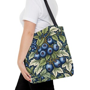 Op de afbeelding: Een zwarte tote bag met een blauw en groen bloemenpatroon met blauwe bessen. De tas heeft zwarte hengsels.