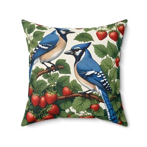 Puede incluir: Una almohada decorativa con un arrendajo azul posado en una rama con fresas rojas. La almohada tiene un fondo blanco con hojas verdes.
