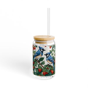 Op de afbeelding: Een herbruikbare glazen tumbler met een bamboe deksel en een doorzichtige rietje. De tumbler heeft een kleurrijk ontwerp met blauwe gaaien en rode aardbeien op een witte achtergrond.