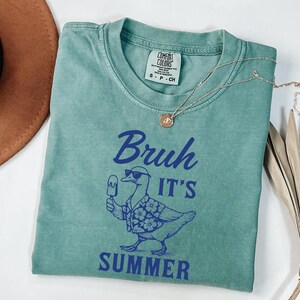 Puede incluir: Una camiseta verde agua con la frase azul "Bruh It's Summer", con un pato con gafas de sol y una camisa hawaiana, sosteniendo un helado. Tambi&eacute;n se ven un collar dorado y un sombrero marr&oacute;n.