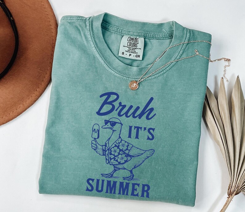 Puede incluir: Una camiseta verde agua con la frase azul "Bruh It's Summer", con un pato con gafas de sol y una camisa hawaiana, sosteniendo un helado. Tambi&eacute;n se ven un collar dorado y un sombrero marr&oacute;n.