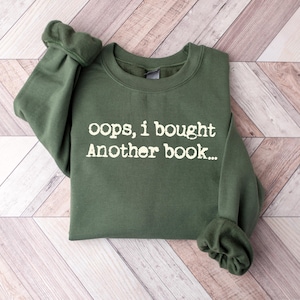 May include: An olive green sweatshirt with the text "oops, i bought Another book..." in white. The sweatshirt is folded on a light brown and white wooden surface. The sleeves are cuffed.