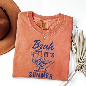 Puede incluir: Una camiseta naranja Comfort Colors con la frase "Bruh It's Summer" en azul, con un pato con gafas de sol y un helado. Tambi&eacute;n hay un collar dorado y un sombrero marr&oacute;n en la imagen.