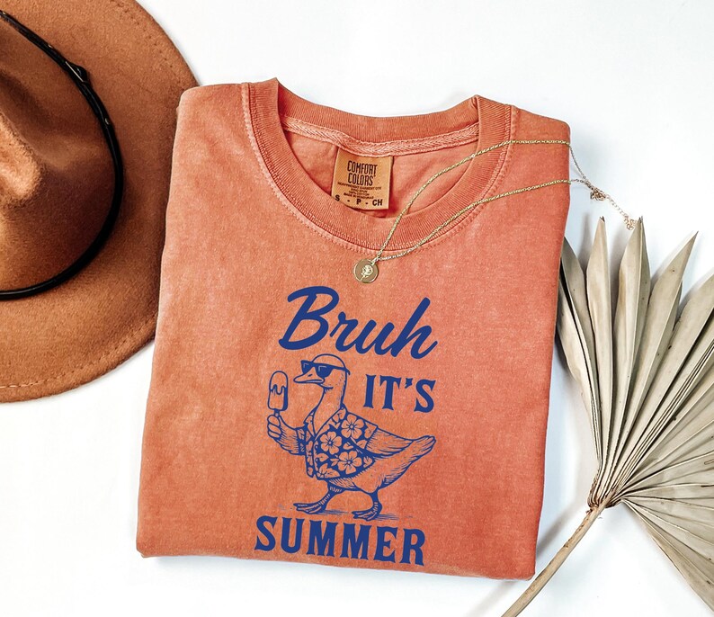 Puede incluir: Una camiseta naranja Comfort Colors con la frase "Bruh It's Summer" en azul, con un pato con gafas de sol y un helado. Tambi&eacute;n hay un collar dorado y un sombrero marr&oacute;n en la imagen.