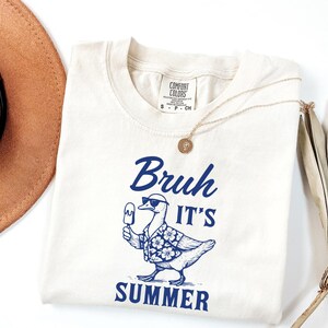 Puede incluir: Camiseta color crema con un gr&aacute;fico azul de un pato con gafas de sol y una camisa hawaiana, sosteniendo un helado. El texto "Bruh It's Summer" est&aacute; encima y debajo del pato. Un collar dorado est&aacute; sobre la camiseta.