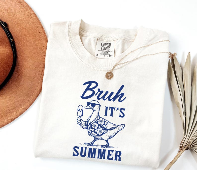 Puede incluir: Camiseta color crema con un gr&aacute;fico azul de un pato con gafas de sol y una camisa hawaiana, sosteniendo un helado. El texto "Bruh It's Summer" est&aacute; encima y debajo del pato. Un collar dorado est&aacute; sobre la camiseta.