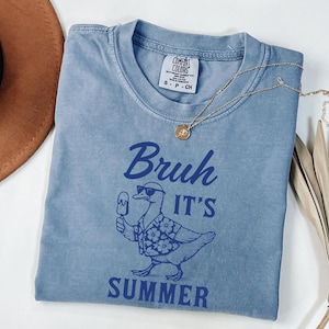 Puede incluir: Una camiseta azul con las palabras "Bruh It's Summer" y un gr&aacute;fico de un pato con gafas de sol y una camisa hawaiana, sosteniendo un helado. Un collar de oro con un colgante est&aacute; sobre la camiseta.