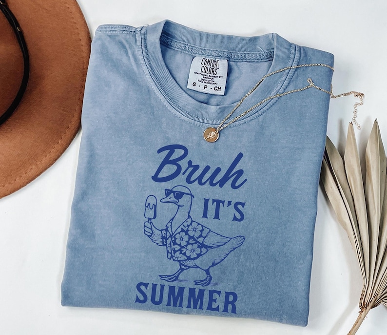 Puede incluir: Una camiseta azul con las palabras "Bruh It's Summer" y un gr&aacute;fico de un pato con gafas de sol y una camisa hawaiana, sosteniendo un helado. Un collar de oro con un colgante est&aacute; sobre la camiseta.