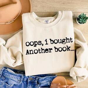 May include: Cream-colored sweatshirt with the text "oops, i bought another book..." in black. The sweatshirt is paired with blue denim shorts, a tan hat, and brown sandals. A small succulent plant is also visible.