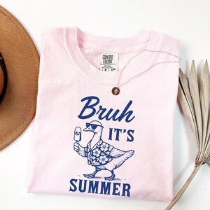 Puede incluir: Una camiseta rosa claro con la frase "Bruh It's Summer" en azul marino. El gr&aacute;fico presenta un pato con gafas de sol y una camisa hawaiana, sosteniendo un helado. Un collar dorado est&aacute; sobre la camiseta.