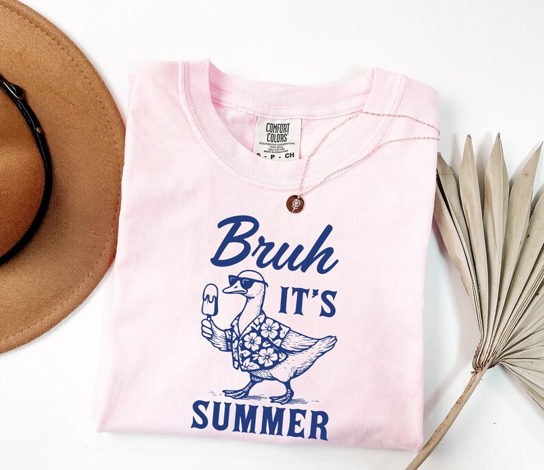 Puede incluir: Una camiseta rosa claro con la frase "Bruh It's Summer" en azul marino. El gr&aacute;fico presenta un pato con gafas de sol y una camisa hawaiana, sosteniendo un helado. Un collar dorado est&aacute; sobre la camiseta.