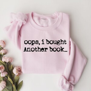 May include: A light pink sweatshirt with the text "oops, i bought Another book..." in black. The sweatshirt is folded and placed on a white surface, with pink flowers and green leaves in the lower left corner.