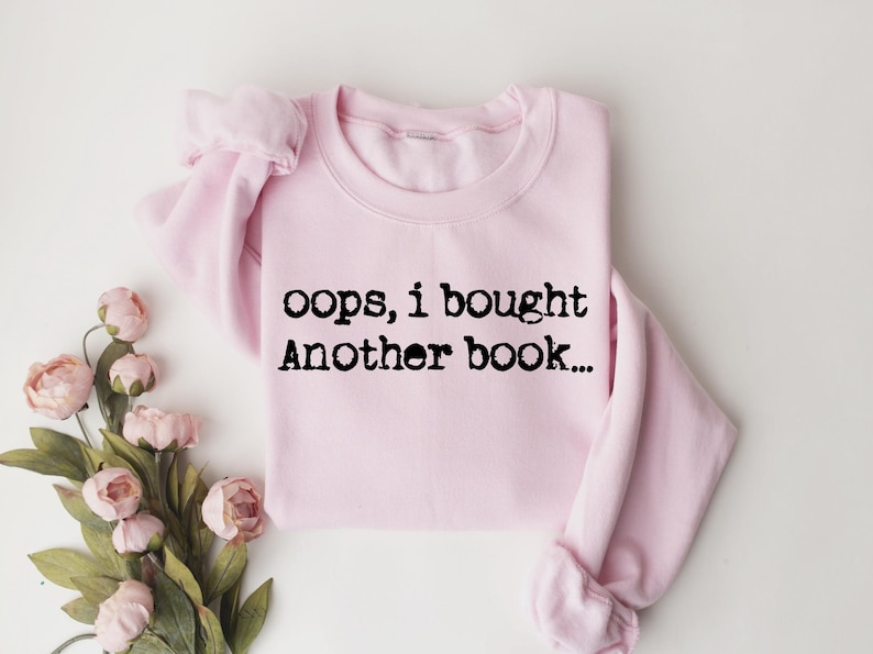 May include: A light pink sweatshirt with the text "oops, i bought Another book..." in black. The sweatshirt is folded and placed on a white surface, with pink flowers and green leaves in the lower left corner.