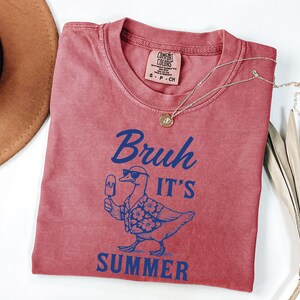 Puede incluir: Una camiseta rojo-naranja con la frase "Bruh It's Summer" en azul. El gr&aacute;fico presenta un pato con gafas de sol y una camisa hawaiana, sosteniendo un helado. Tambi&eacute;n se ve un collar dorado y un sombrero marr&oacute;n.