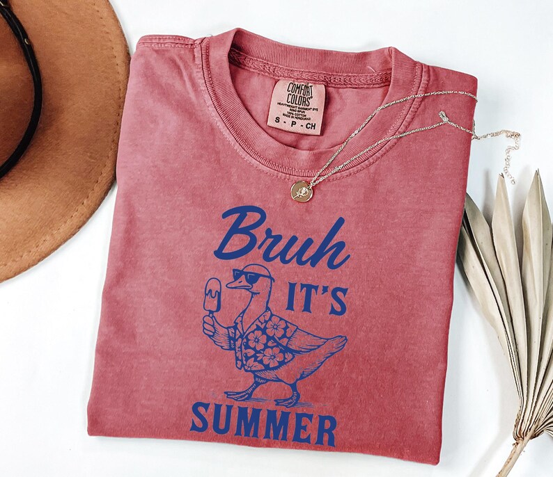 Puede incluir: Una camiseta rojo-naranja con la frase "Bruh It's Summer" en azul. El gr&aacute;fico presenta un pato con gafas de sol y una camisa hawaiana, sosteniendo un helado. Tambi&eacute;n se ve un collar dorado y un sombrero marr&oacute;n.