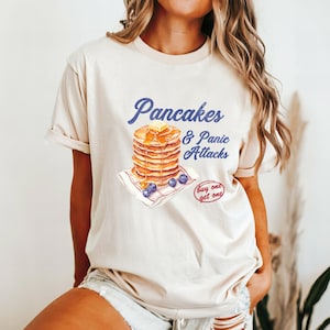 May include: Cream-colored t-shirt with the text "Pancakes & Panic Attacks" in blue script. A stack of pancakes with butter and syrup is depicted, along with blueberries. A small "buy one get one" graphic is also visible.