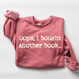 May include: A red-pink sweatshirt with the text "oops, i bought Another book..." in white lettering. The long-sleeved shirt is folded, with the cuffs of the sleeves rolled up.