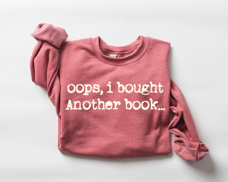 May include: A red-pink sweatshirt with the text "oops, i bought Another book..." in white lettering. The long-sleeved shirt is folded, with the cuffs of the sleeves rolled up.