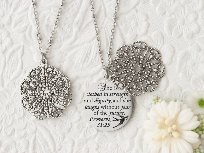May include: Two silver-toned necklaces with intricate, openwork floral pendants. One pendant is round with the text "She is clothed in strength and dignity, and she laughs without fear of the future. Proverbs 31:25".
