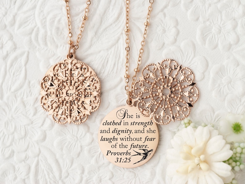 May include: Rose gold necklaces with intricate filigree pendants. One necklace features a round pendant with the text "She is clothed in strength and dignity, and she laughs without fear of the future. Proverbs 31:25" and a small bird.
