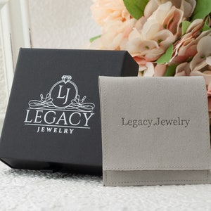 May include: A black jewelry box with the Legacy Jewelry logo and a gray pouch with "Legacy.Jewelry" printed on it. The box is open, and the pouch is folded. The background includes flowers and a white framed object.