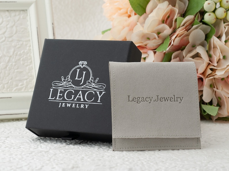 May include: A black jewelry box with the Legacy Jewelry logo and a gray pouch with "Legacy.Jewelry" printed on it. The box is open, and the pouch is folded. The background includes flowers and a white framed object.