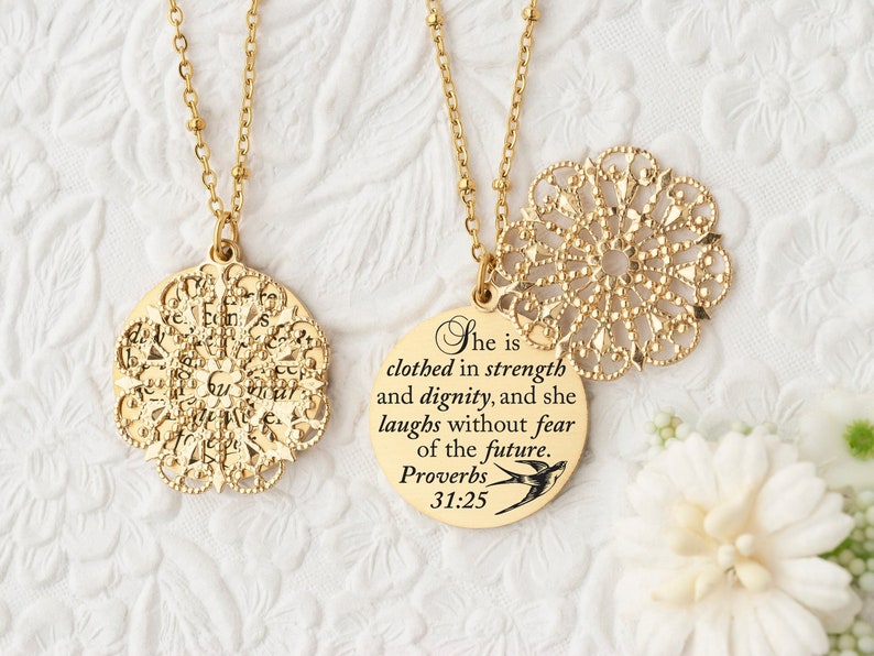 May include: Two gold-colored necklaces with round pendants. One pendant has a detailed openwork design, the other has the text "She is clothed in strength and dignity, and she laughs without fear of the future. Proverbs 31:25."