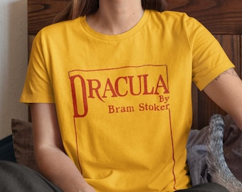 Könnte beinhalten: Eine Frau trägt ein gelbes T-Shirt mit dem Text "Dracula by Bram Stoker" in einem roten Kasten. Sie sitzt auf einem Bett mit einer weiß-grauen Katze.