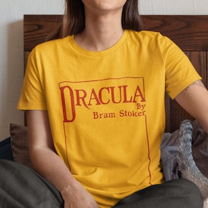 May include: A woman wearing a yellow t-shirt with the text "Dracula by Bram Stoker" in a red box. She is sitting on a bed with a white and gray cat.