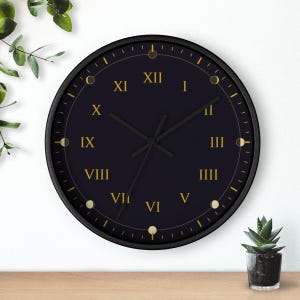 May include: A round wall clock with a black face and a black frame. The clock features gold Roman numerals from I to XII and gold hands. Small gold circular icons representing moon phases are positioned at the hour markers around the dial.