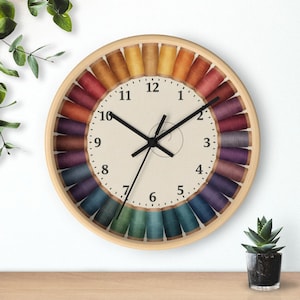 May include: A round wall clock with a light wood frame featuring a cream-colored face with black numerals from 1 to 12. The perimeter is decorated with a circular arrangement of colorful thread spools in a gradient of yellow, red, purple, and blue.