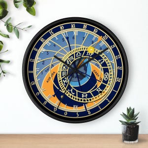 Op de afbeelding: Een ronde wandklok met een astronomisch wijzerplaatontwerp en een zwarte rand. De wijzerplaat heeft een blauw en oranje kleurenschema met Romeinse cijfers, dierenriemsymbolen en de tekst ORTUS, AURORA en CREPUSCULUM in gouden letters.