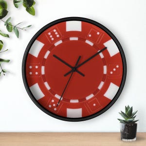 May include: A round wall clock with a black frame and a red face designed to resemble a casino poker chip. The face features white rectangular accents around the perimeter and small white dice patterns with varying dot counts, plus three black hands.