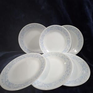 May include: Six white ceramic plates with a light blue floral border. The plates are displayed on a dark blue surface. Each plate has a simple, elegant design, perfect for everyday use. The plates are approximately 20 cm in diameter.