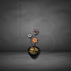 May include: A unique floral arrangement featuring three metal flowers in a black pot filled with brass bullet casings. The flowers are silver, copper, and bronze, with brown stems. The pot is set against a gray background.