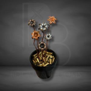 May include: A black ceramic pot filled with brass bullet casings, with metal flowers made from bullet casings. The flowers have copper, silver, and bronze petals. The stems are brown. The background is a gradient of gray.