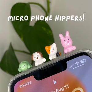 May include: Four small animal-shaped phone accessories labeled MICRO PHONE HIPPERS! include a green turtle, a brown and white dog, a white and orange cat, and a pink rabbit, all attached to the top edge of a smartphone screen.