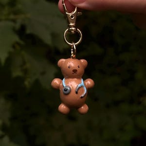 May include: A brown teddy bear keyring featuring a light blue stethoscope draped around its neck. The bear has a smooth, glossy finish, black eyes, and a small black nose. It is attached to a gold-toned metal lobster clasp and ring.