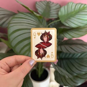 May include: A playing card featuring the Queen of Hearts, with a stylized illustration of a woman in a red and gold outfit. The card has a cream-colored background with gold trim and the letter "Q" and heart symbols.