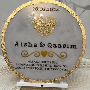 Puede incluir: Una placa circular blanca con un borde dorado y detalles de pan de oro. La placa presenta un diseño en forma de corazón con caligrafía árabe en oro, los nombres "Aisha & Qaasim" en negro y la fecha "26.02.2024" en negro. La placa también incluye el texto "MAY ALLAH BLESS YOU, AND SHOWER HIS BLESSING UPON YOU AND JOIN YOU TOGETHER IN GOODNESS" en negro.