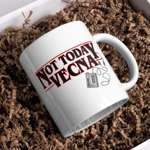 May include: White ceramic mug with the text "NOT TODAY VECNA" in red and black lettering. A black and white illustration of a cassette player with headphones is also on the mug. The mug is on a bed of brown packing material.