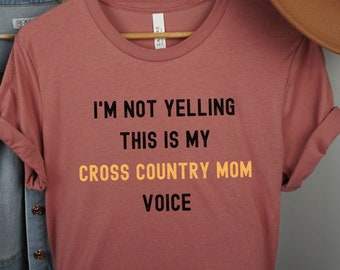 Könnte beinhalten: Ein staubiges rosafarbenes T-Shirt mit dem Text "I'm not yelling this is my cross country mom voice" in Schwarz und Orange.